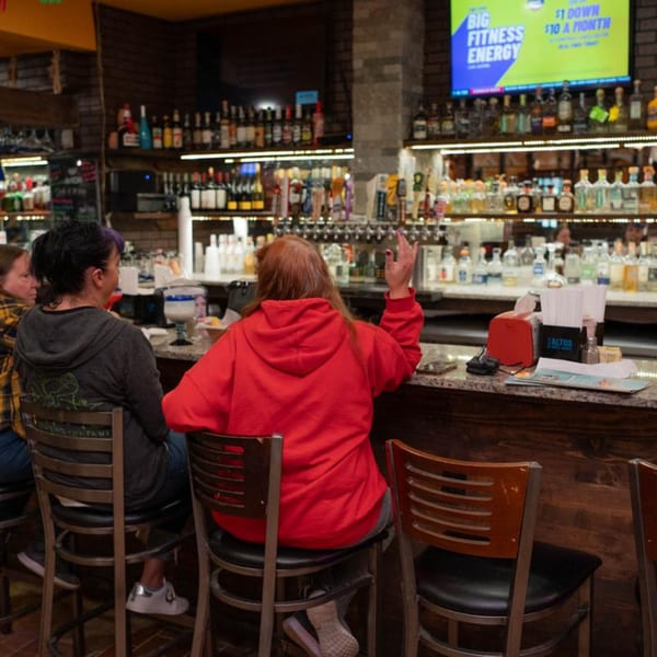 Customers seated at the bar of Papa's & Beer, with bottles of liquor and a TV screen in the background.