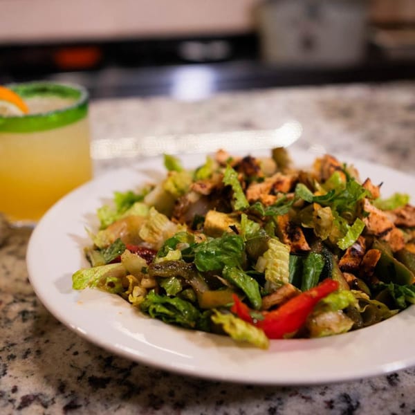 A vibrant Mexican salad with grilled chicken and vegetables, alongside a margarita with a lime garnish.