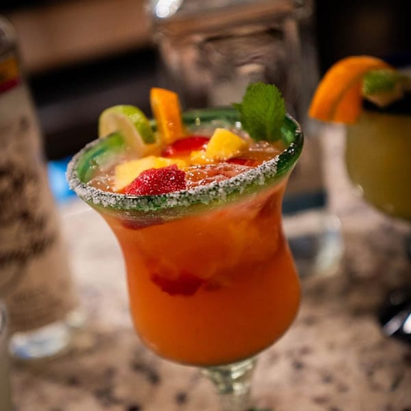 A vibrant fruit margarita with a salted rim, garnished with lime, orange, mint, strawberries, and pineapple.